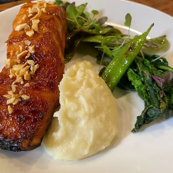 Glazed salmon with chopped nuts, mashed potatoes, and green vegetables on a white plate.
