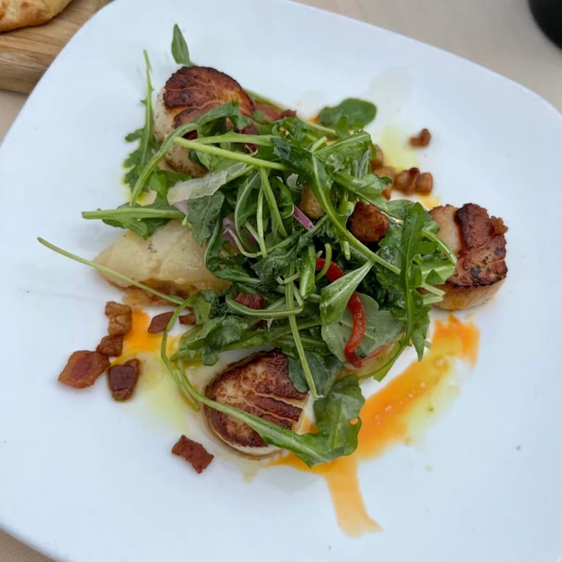 Seared scallops with arugula, bacon bits, and a vibrant orange sauce on a white plate.