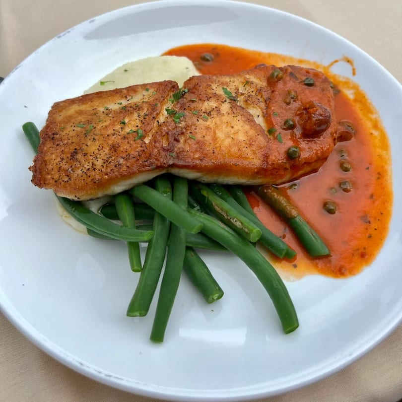 Pan-seared fish with red sauce, capers, mashed potatoes, and green beans on a white plate.